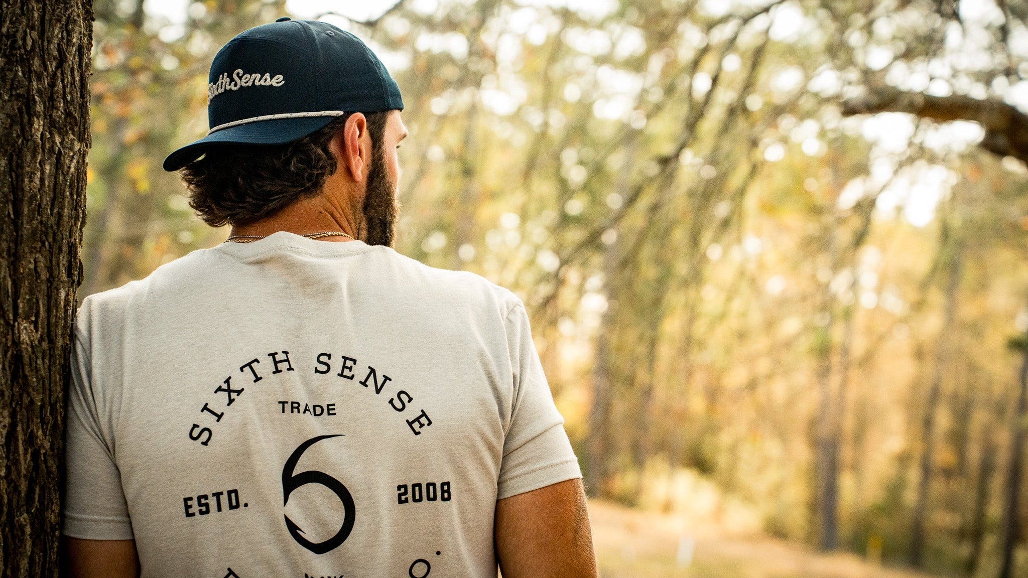 Person wearing a cap and t-shirt with 'Sixth Sense' branding in a forest setting